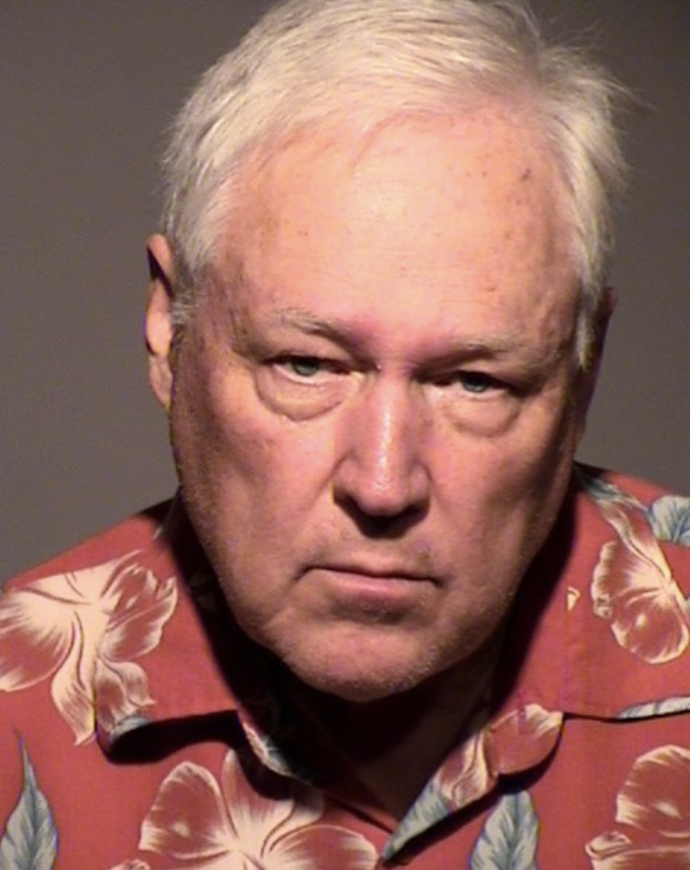 Mugshot of white man, older, white hair, growning, wearing red hawaiaan shirt.